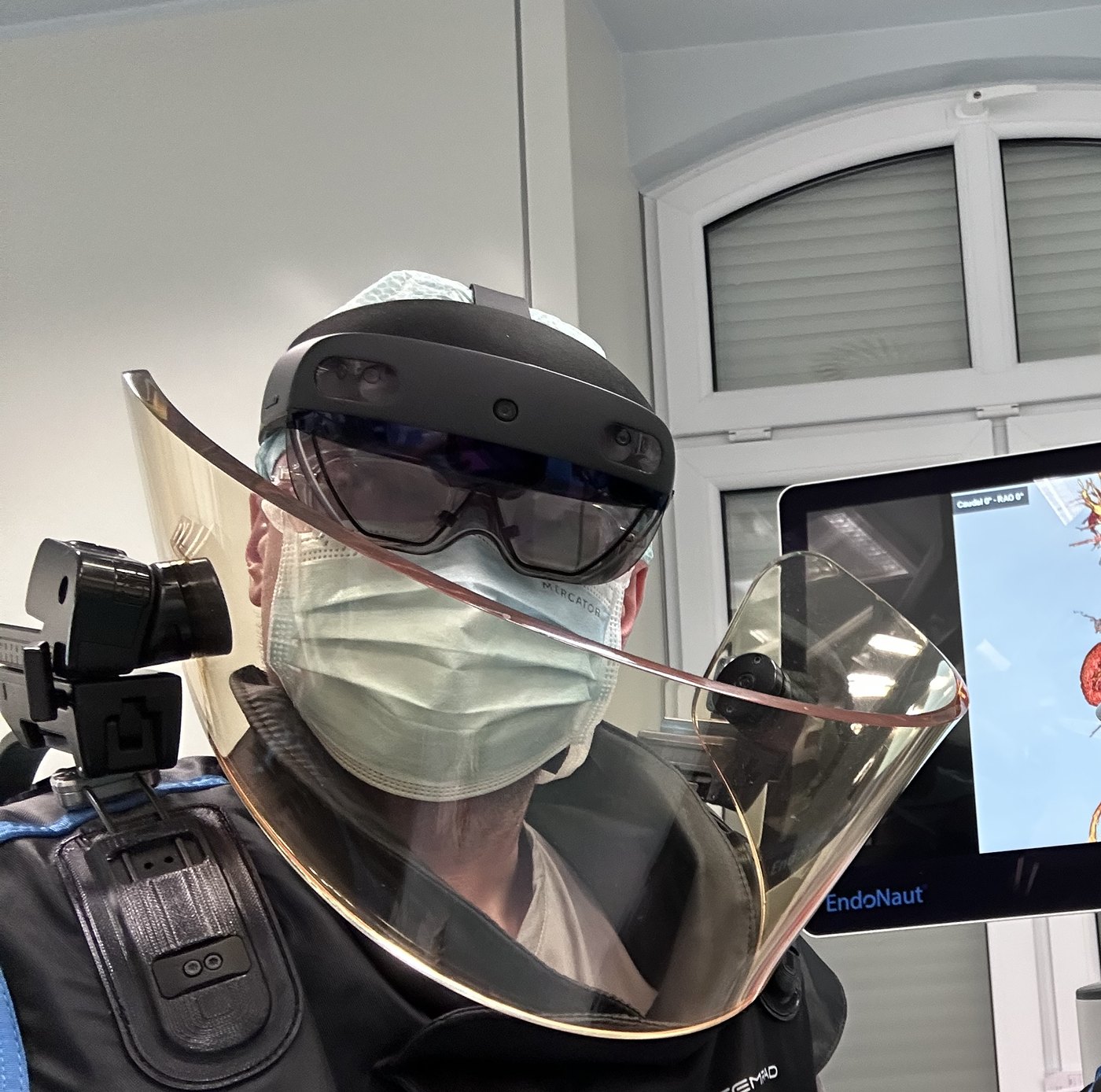 dr hab. Paweł Rynio w goglach mieszanej rzeczywistości HoloLens i egzoszkielecie radiacyjnym na sali zabiegowej; w tle monitor z trójwymiarowym modelem aorty